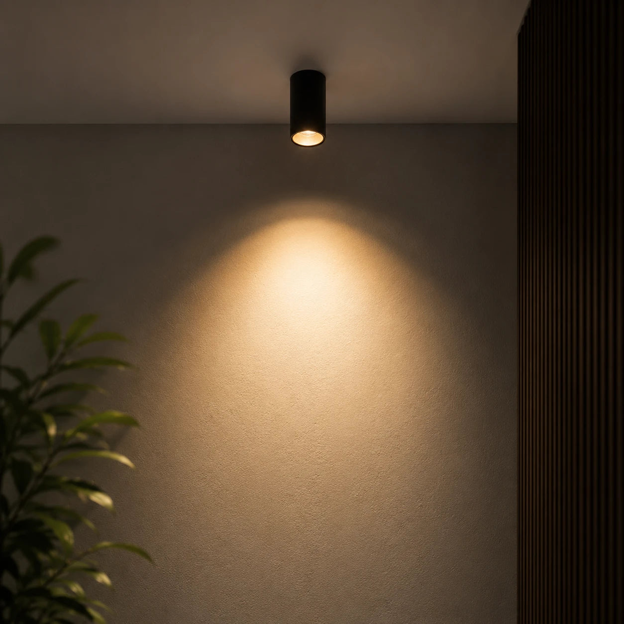 A modern black cylindrical ceiling spotlight mounted on a textured beige wall, casting a warm yellow light downward, with a dark slatted panel on the right and a green plant on the left.