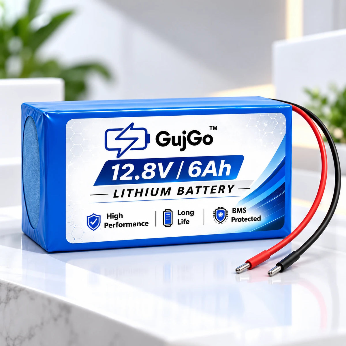 A blue and white GujGo 12.8V 6Ah lithium battery with red and black cable leads, displayed on a white marble surface with a blurred indoor background.