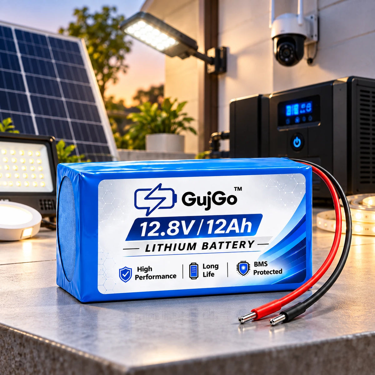 A blue and white GujGo 12.8V 12Ah lithium battery with attached red and black cables, displayed on a metal workbench with solar panels, lights, and a power unit in the background.