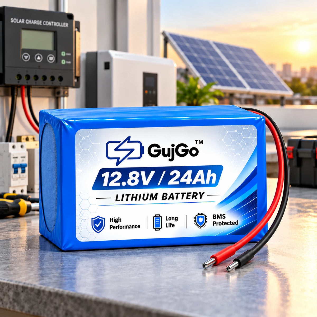A blue and white GujGo 12.8V/24Ah lithium battery in a rectangular metal case, shown on a workbench with solar panels and a solar charge controller in the background.