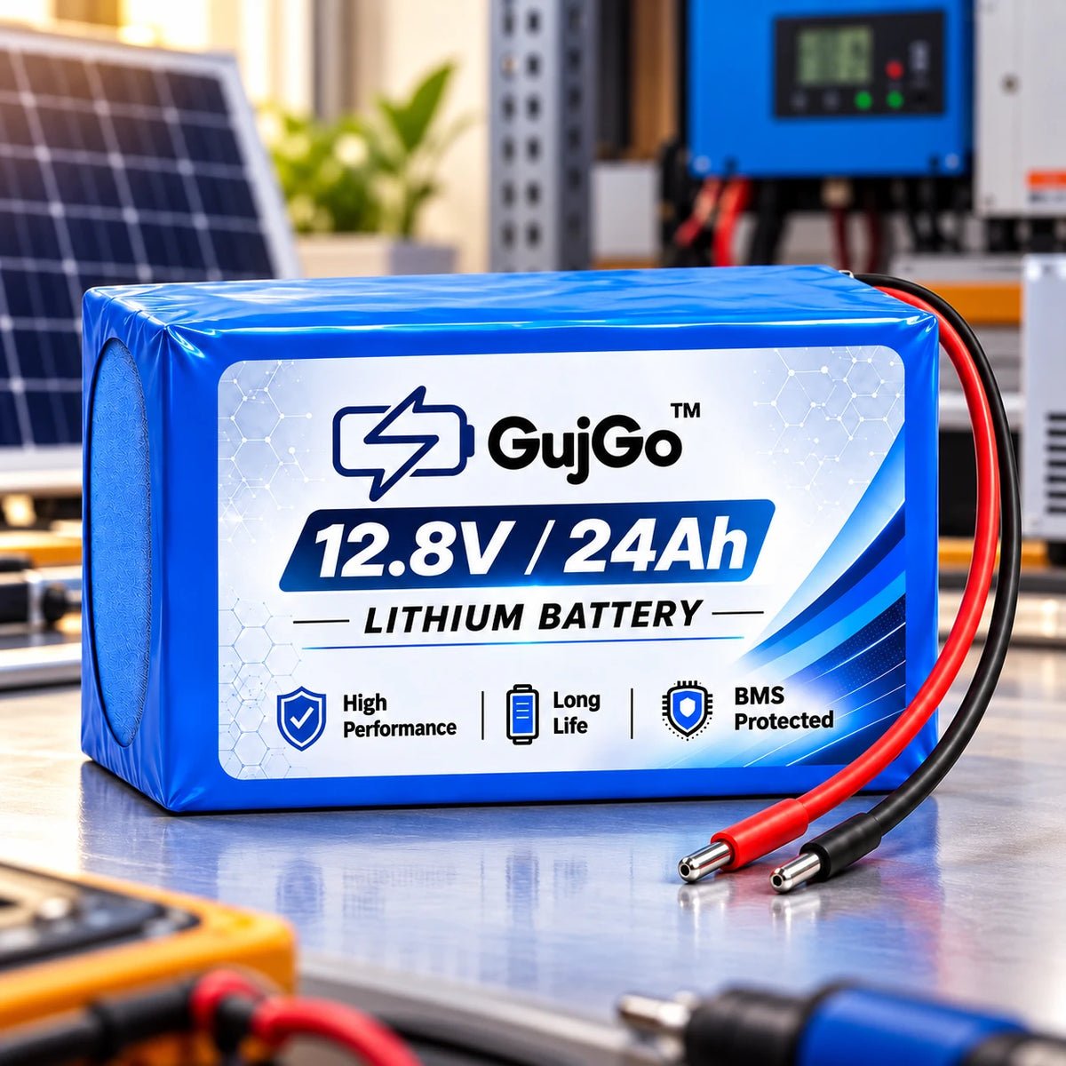 A blue and white GujGo 12.8V / 24Ah lithium battery pack with attached red and black cable leads, displayed on a workbench with a blurred background of electronic tools and a solar panel.