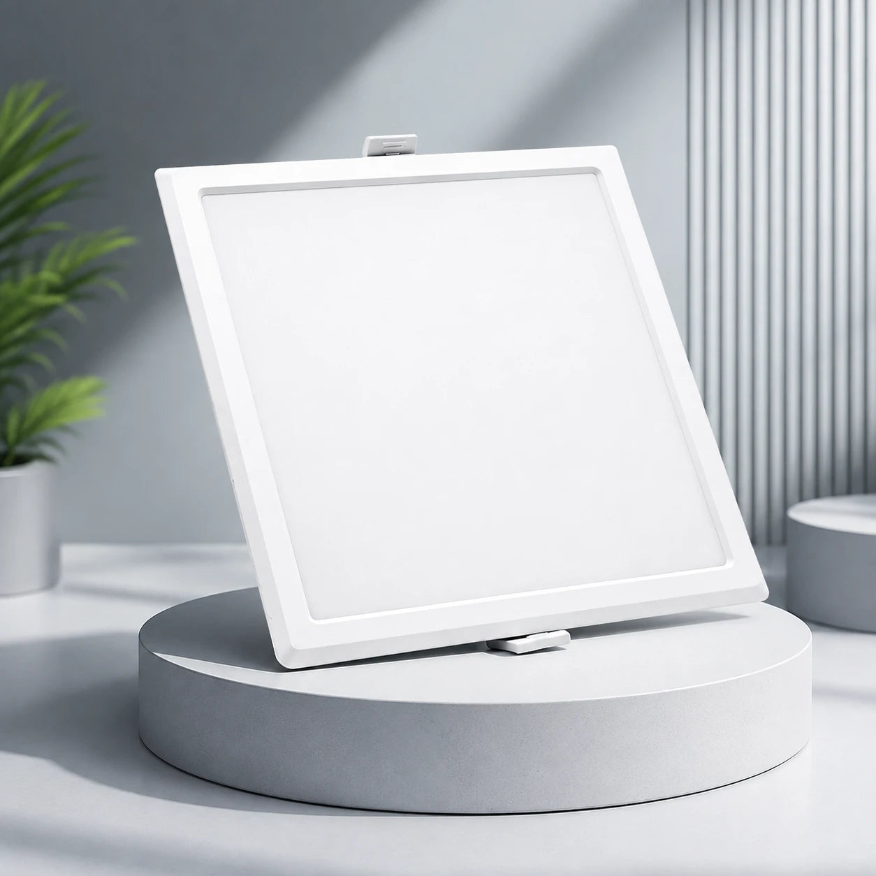 A square white LED ceiling light panel shown at an angle on a round pedestal, with a modern gray background and a small potted plant in the side.