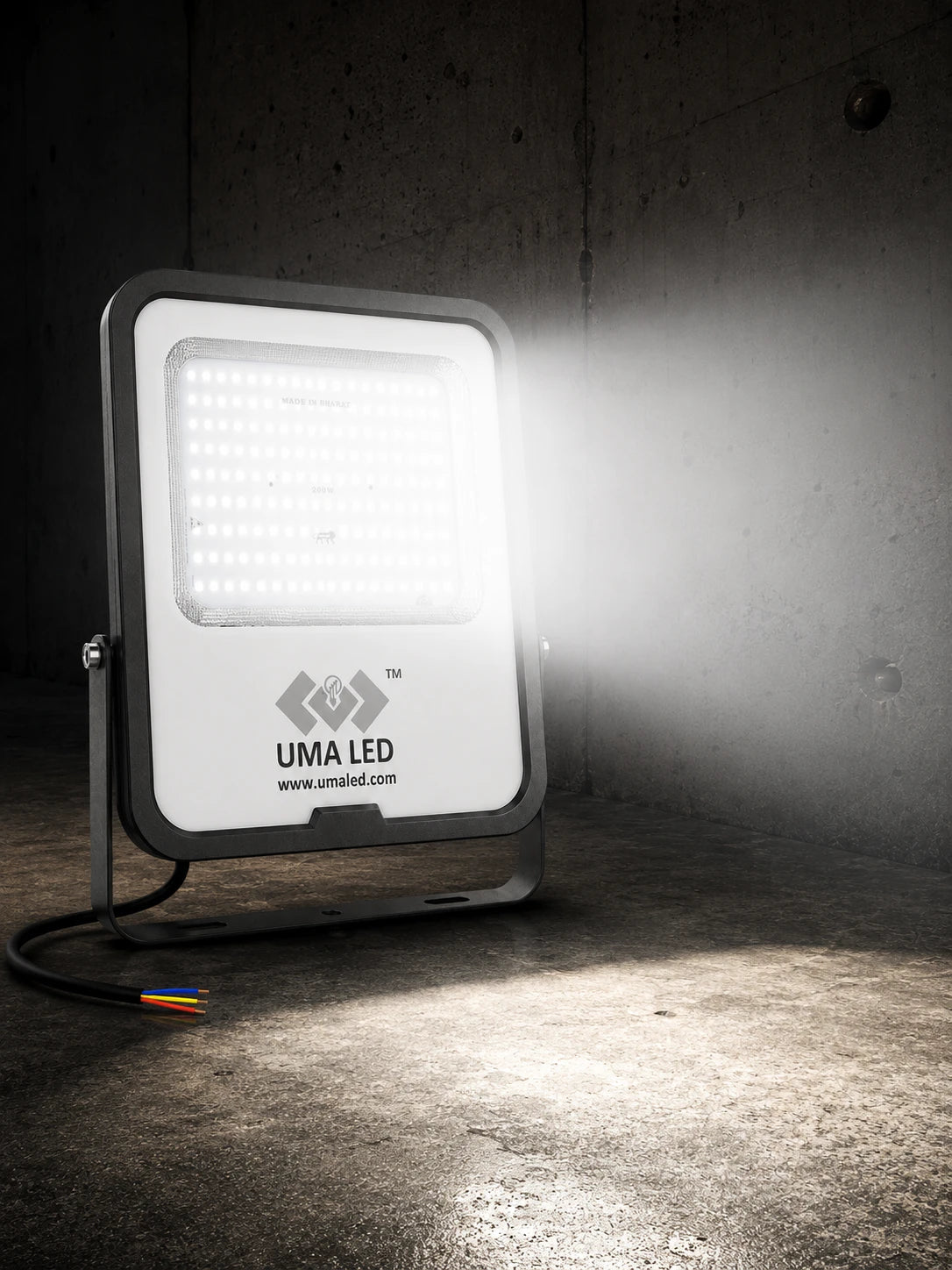 A UMA LED floodlight with a white faceplate and dark gray housing, shown turned on and casting a bright beam of light onto a concrete wall and floor.