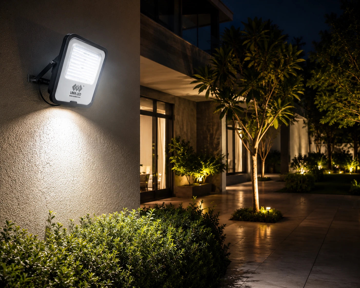 A modern outdoor wall-mounted LED light fixture illuminating a stucco exterior wall at night, with landscaped shrubs, trees, and a walkway in the background.