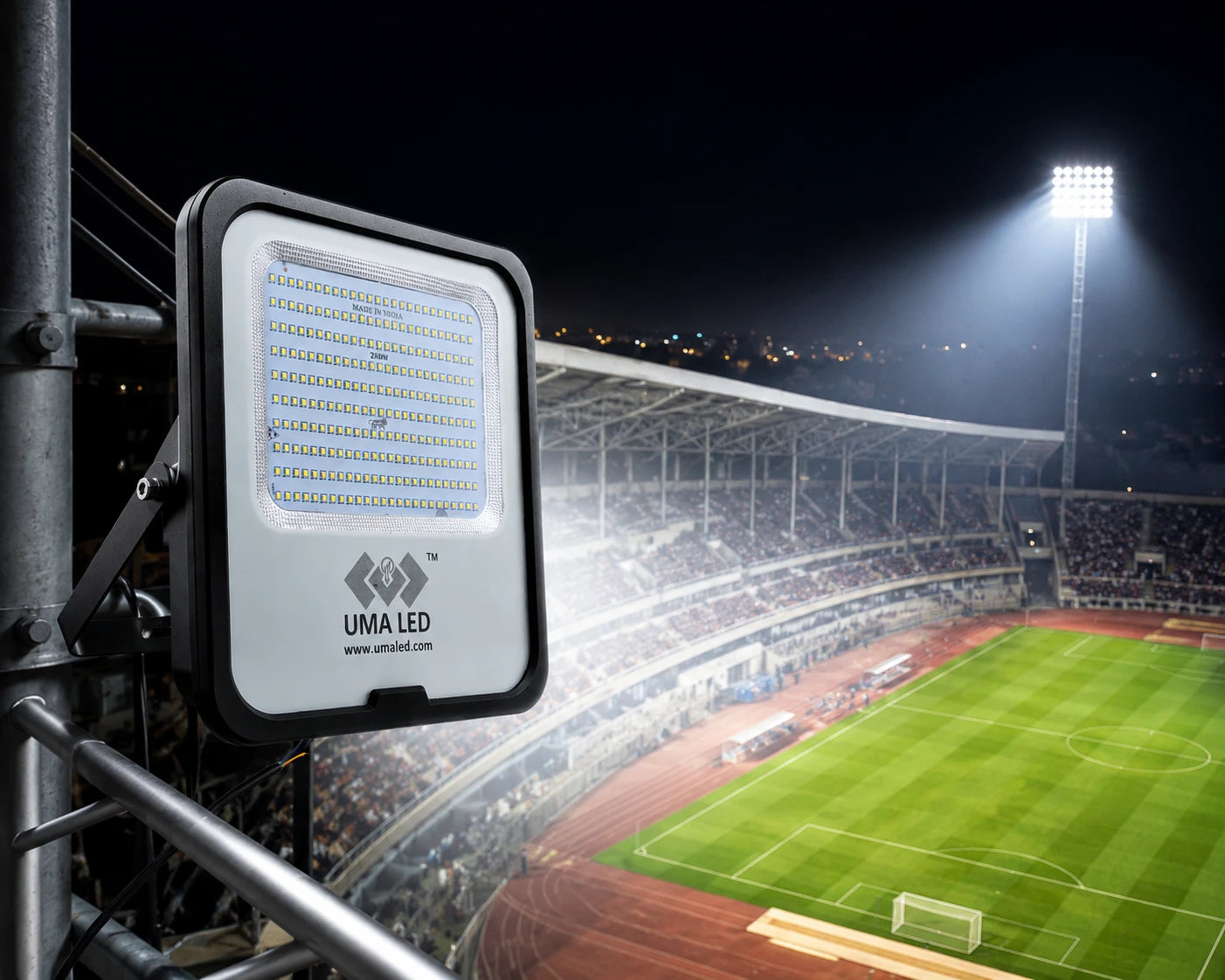 A large rectangular UMA LED floodlight mounted on a stadium structure, shown in the foreground with a brightly lit soccer field and stands in the background at night.