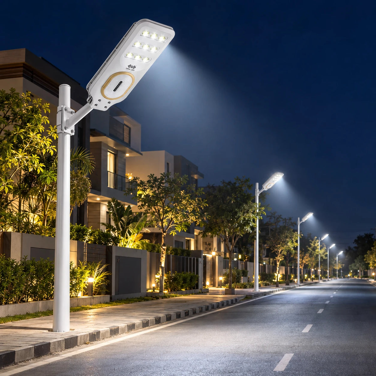 A modern white solar street light mounted on a tall pole, illuminating a quiet residential road at night. The light has a rectangular LED panel with a circular gold accent and is shown in a row of other matching lights along a landscaped street with houses and trees in the background.