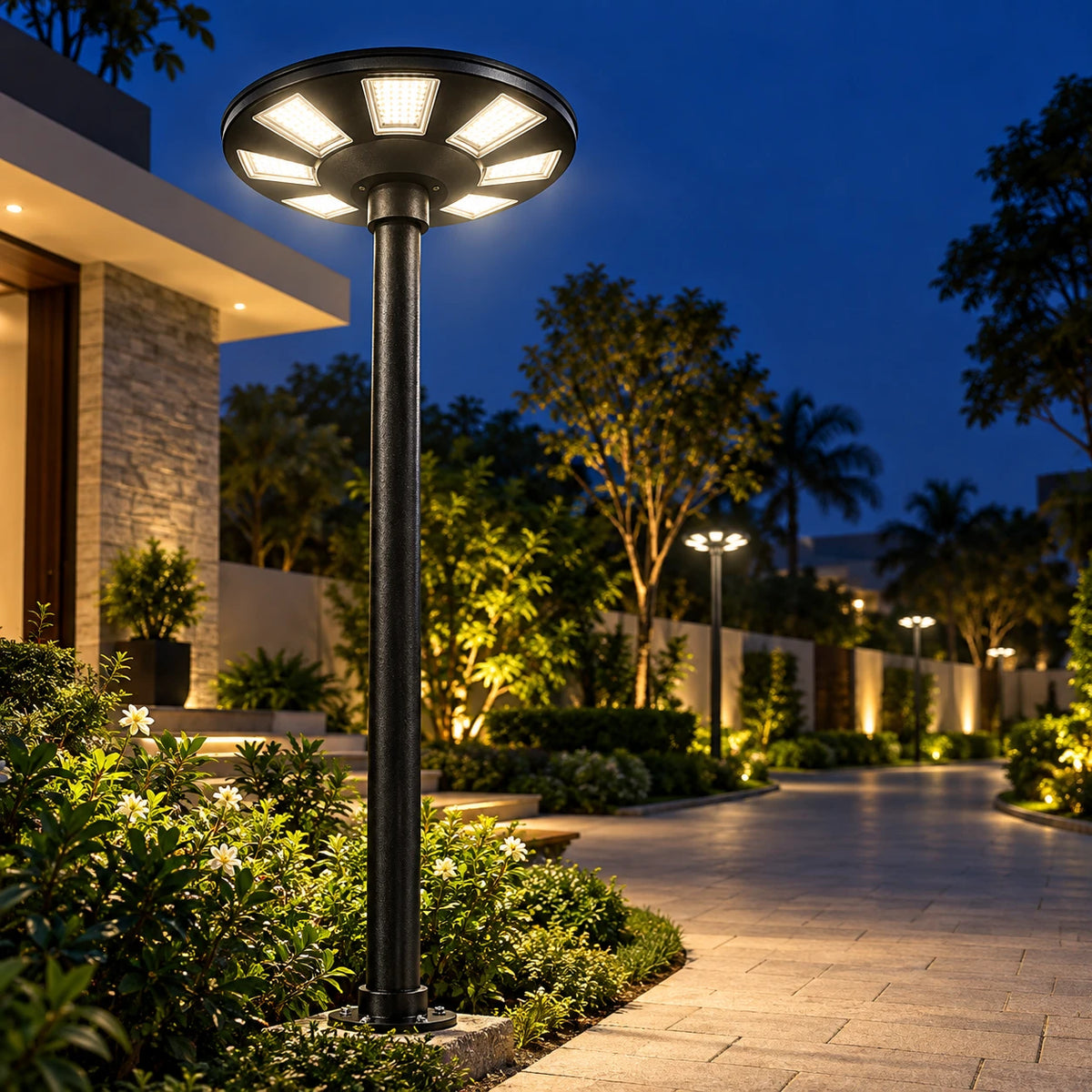 A modern black outdoor bollard light post with a circular multi-light head, illuminated at dusk in a landscaped residential courtyard with a paved walkway and greenery.