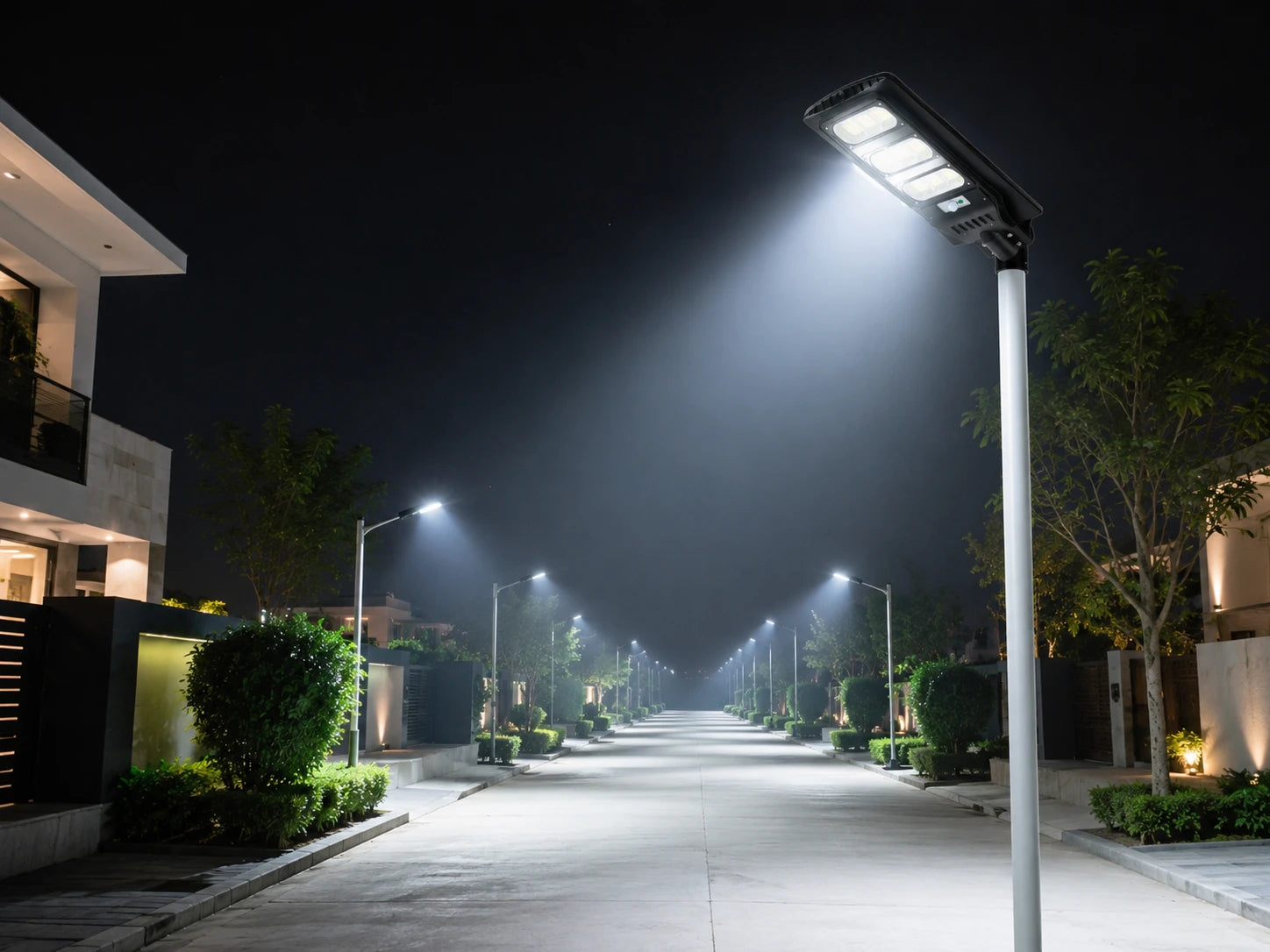 A modern solar street light mounted on a tall pole illuminates a residential pathway at night, with multiple matching lights lining the road between houses and trees.